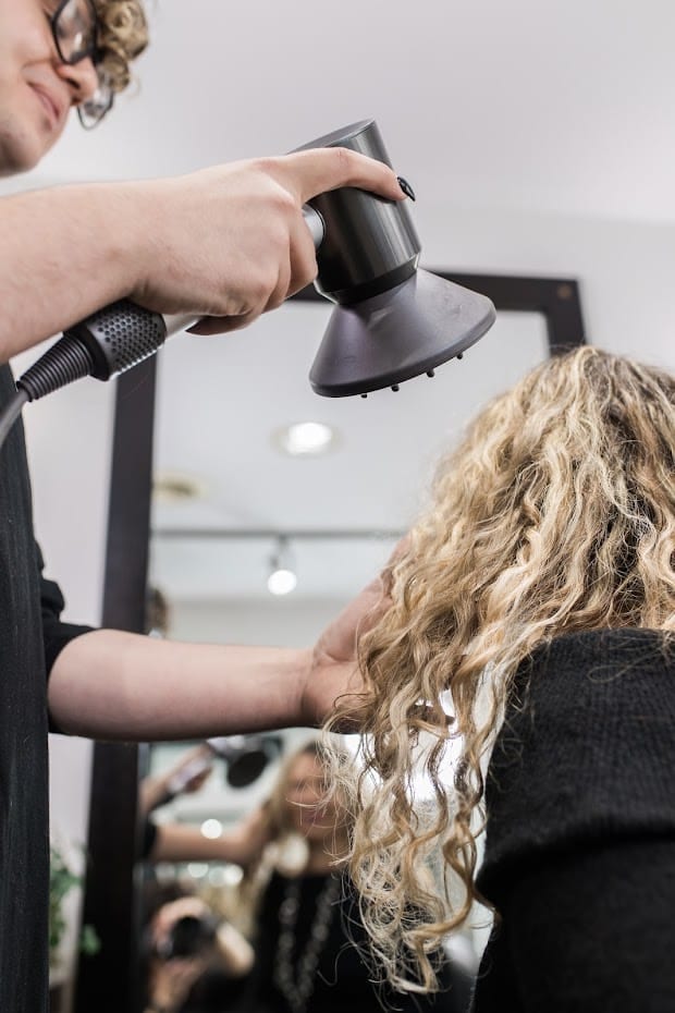 A stylist at YOLOve Beauty Spa in Dartmouth uses a hairdryer diffuser to style curly blonde hair. The client, seated and facing a mirror, enjoys the bright, modern salon ambiance.
