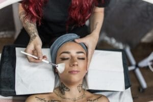 At YOLOve Beauty Spa in Dartmouth, a client with tattoos receives a facial treatment from a red-haired beautician while lying on a treatment bed, wearing a blue headband.